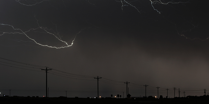 10 wooden poles on an overhead, 3-phase feeder at night in a rural area. Two large strokes of lightning and dark clouds line the sky above the feeder